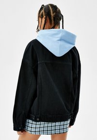 Svart denimjacka med en ljusblå huva, lös passform, synlig sömnad och knappar. Bakgrunden är helt vit.