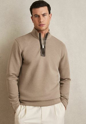 Sudadera con cremallera de color marrón claro y cuello alto, con una cremallera de color marrón oscuro y una textura suave. El modelo lleva pantalones blancos.