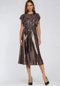 Kobieta w błyszczącej metalicznej sukience midi z falbankowymi rękawami i przewiązanym paskiem w talii, zestawiona z czarnymi szpilkami, stoi na jednolitym tle.