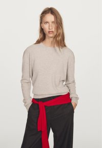 Beige stickad tröja med rund halsringning och långa ärmar, tillsammans med lösa svarta byxor som har ett rött tygbälte knutet i midjan.