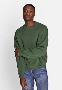 Homme noir aux cheveux courts portant un pull vert et un jean bleu, debout devant un fond gris clair uni, bras croisés.