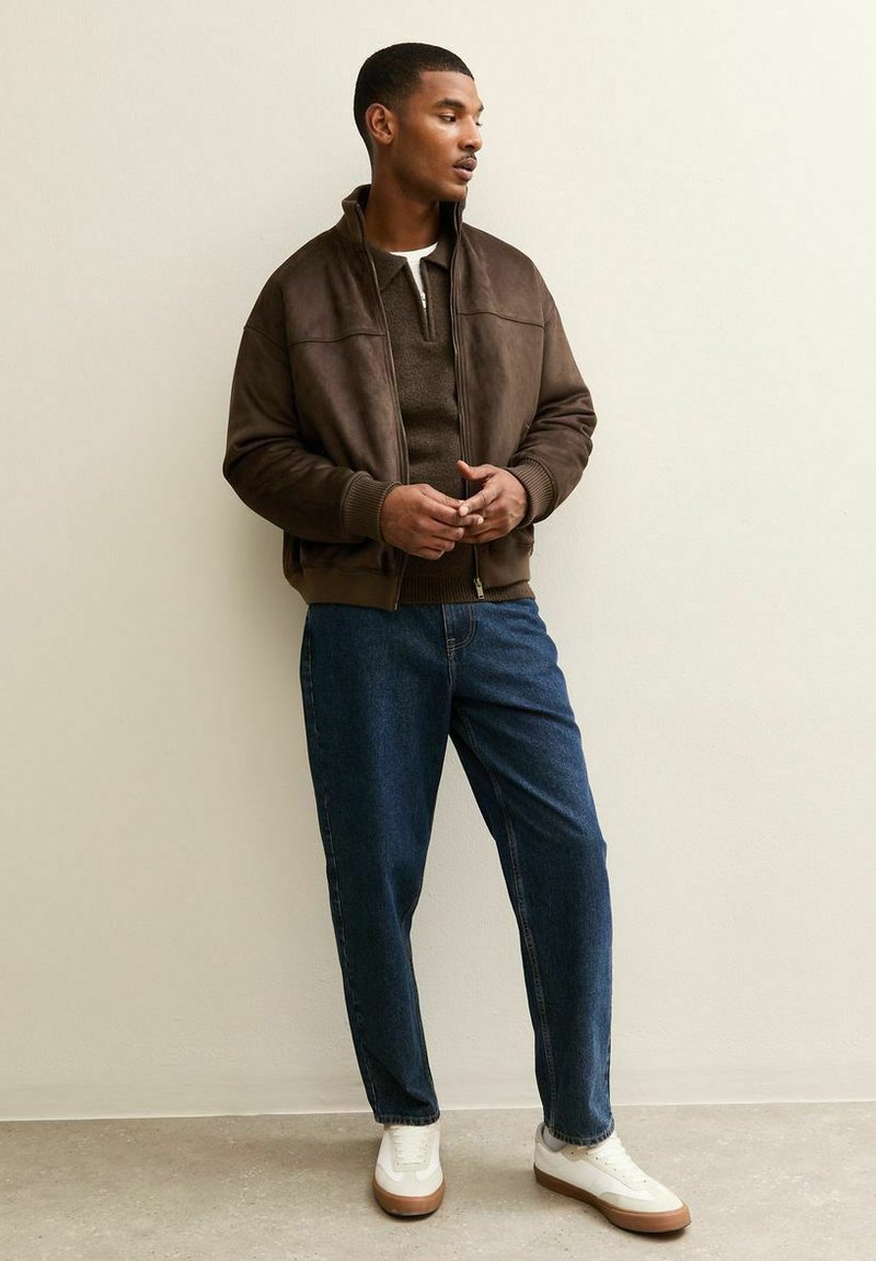 Bruine leren bomberjack met een hoge kraag, gedragen over een donker trui, gecombineerd met blauwe spijkerbroeken en witte sneakers.