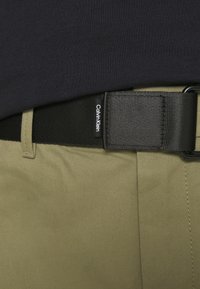 Zwarte gevlochten riem met een rechthoekige gesp, voorzien van een "Calvin Klein" label. Draag een licht olijfgroene katoenen broek.