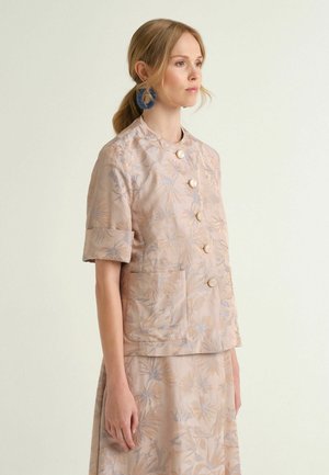 Frau mit hellem Haar in einem niedrigen Pferdeschwanz, die eine beige Jacke mit floralem Muster und großen Knöpfen sowie blaue Creolen trägt, seitlich stehend.