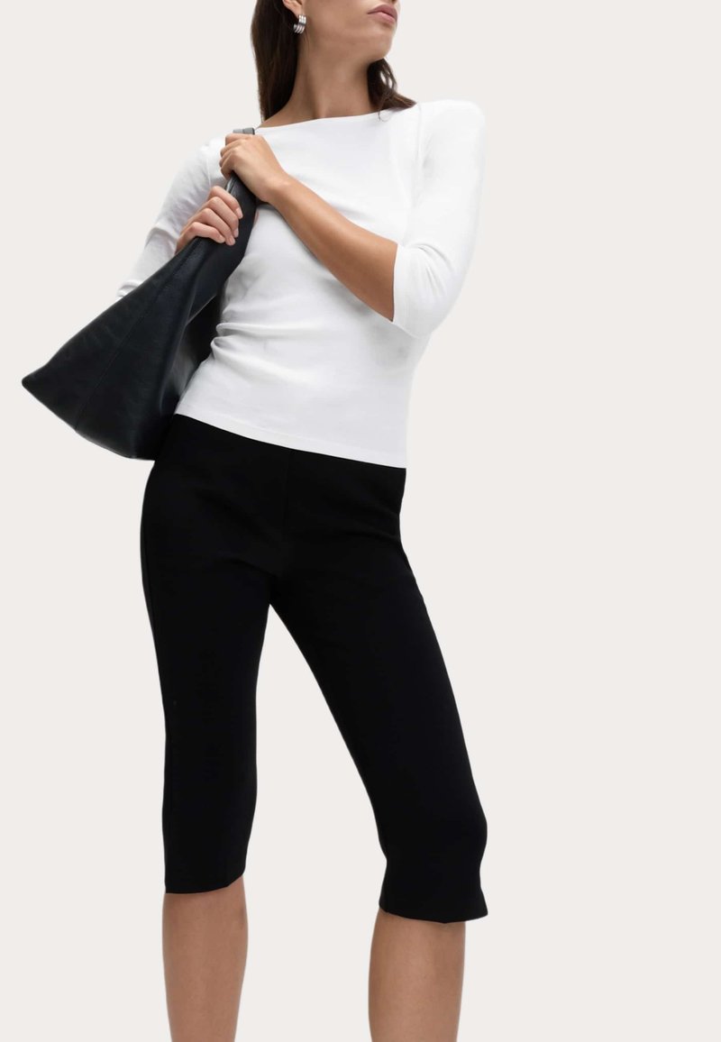 Femme portant un haut blanc à manches trois-quarts et des leggings noirs jusqu'aux genoux, tenant un grand sac à main noir sur l'épaule.