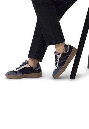 Baskets en daim bleu et marine avec une touche en cuir blanc, semelle en caoutchouc texturé et détails cousus, associées à un pantalon sombre.
