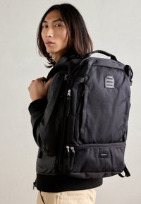 Black backpack with a textured finish, multiple zippered compartments, hexagonal logo detail, padded straps, and a structured shape.