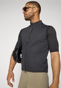 Gilet de cyclisme noir à manches courtes, zip à l'avant et matériau texturé. Le modèle tient un casque noir. Les pantalons sont de couleur beige clair.