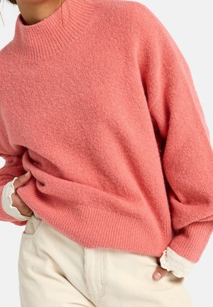 Pull rose doux avec col montant et poignets côtelés, porté sur un pantalon en velours côtelé couleur crème, les mains posées négligemment dans les poches.