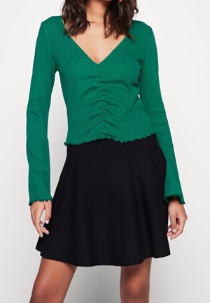 Vrouw draagt een groene lange mouwen top met V-hals en plooien, gecombineerd met een zwarte A-lijn rok tegen een witte achtergrond.