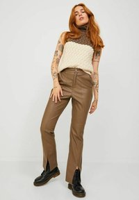 Suéter de cuello alto trenzado marrón sobre un panel texturizado color crema; pantalones de imitación de cuero marrón con aberturas laterales; botas negras con cordones y suela amarilla.