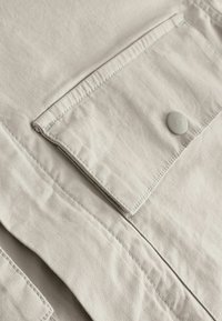 Beiger Baumwollstoff mit Knopfverschluss und einer aufgesetzten Tasche. Verfügt über kontrastierende Nähte und eine weiche Textur.