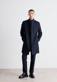 Lungo cappotto blu navy scuro con colletto alto e chiusura a bottone, indossato sopra un completo coordinato, abbinato a scarpe nere, su uno sfondo neutro.