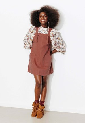 Jeune femme avec une grande coiffure afro portant un chemisier fleuri, une robe tablier couleur rouille, des chaussettes bordeaux et des bottes mocassins marron, souriant et posant.
