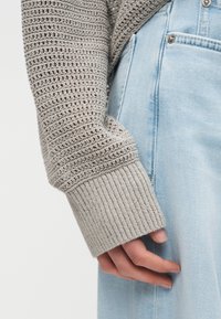 Main dans une manche de pull tricoté gris clair reposant contre un jean en denim bleu clair avec une couture visible et une fermeture à bouton argenté.