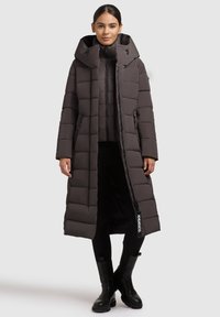 Donna in piedi con un lungo cappotto imbottito marrone scuro dotato di un ampio cappuccio, leggings neri e stivaletti neri su uno sfondo neutro.