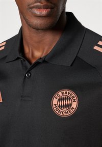 Musthave must jälgimisriidega, millel on tekstuur, roosakuldne FC Bayern Müncheni logo ja kolm roosakuldset triipu vasakul õlal.