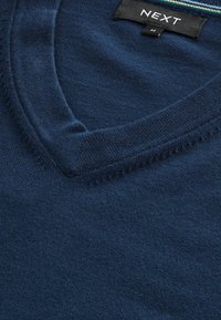 Pull en tricot bleu marine avec un col en V, une texture lisse, et une étiquette marquée "NEXT" avec un accent rayé contrastant.