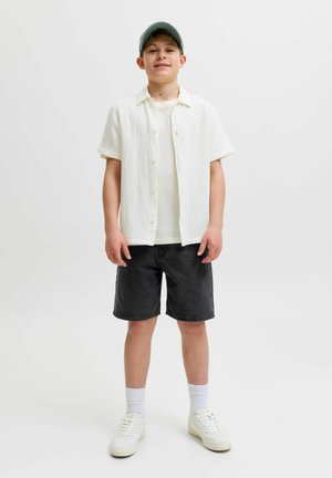 Dječak obučen u bijelu košulju kratkih rukava preko bijele majice, crne kratke hlače, bijele čarape, tenisice i zelenu baseball kapu, stoji ispred bijele pozadine.