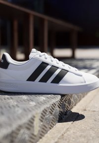 Baskets blanches Adidas avec des bandes noires posées sur une rampe métallique texturée en plein air en pleine journée.