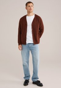 Bruine gebreide cardigan met een V-hals, knoopsluiting aan de voorkant en zijzakken, gedragen over een wit T-shirt en gecombineerd met lichtblauwe jeans.