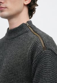 Mann mit einem dunkelgrauen, strukturierten Strickpullover mit einem goldenen Reißverschluss, der vom Kragen bis zur Schulternaht verläuft.