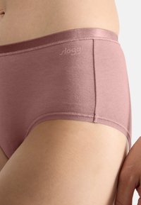 Slip in cotone rosa con texture liscia, caratterizzato da un design senza cuciture e un logo sul cinturino. Colore discreto e stile minimalista.