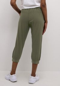 Personne portant un pantalon court olive à poignets élastiques et des baskets blanches, debout devant un fond clair uni.