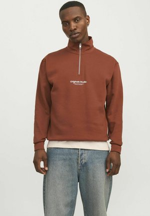 Brauner Reißverschluss-Sweatshirt mit hohem Kragen, vorne mit einem weißen "Originals Studio"-Logo versehen, kombiniert mit hellblauen Jeans.