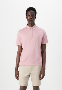 Ανοιχτό ροζ πόλο πουκάμισο από μαλακό βαμβάκι με γιακά, τρία κουμπιά και μικρό λογότυπο στην αριστερή πλευρά του στήθους. Συνδυασμένο με μπεζ σορτς.