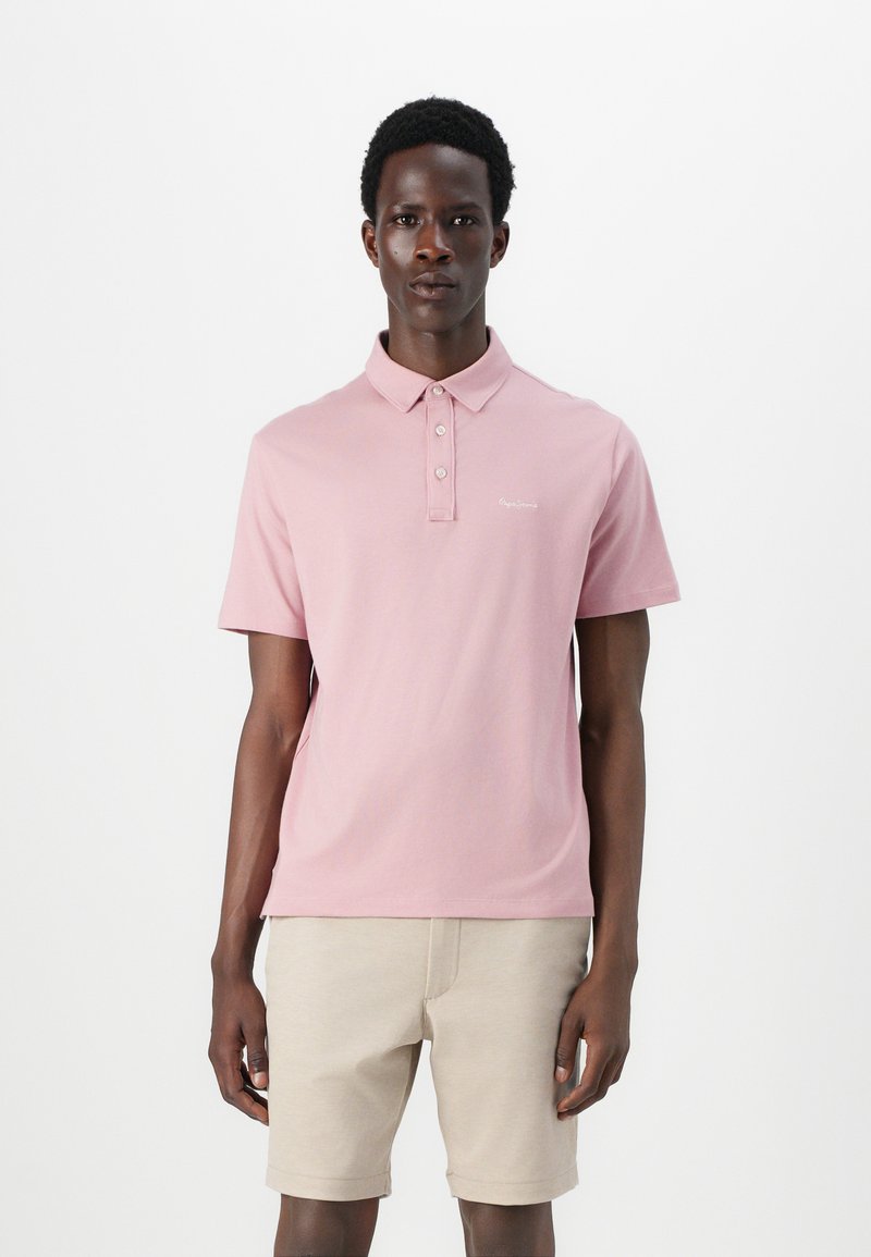 Ανοιχτό ροζ πόλο πουκάμισο από μαλακό βαμβάκι με γιακά, τρία κουμπιά και μικρό λογότυπο στην αριστερή πλευρά του στήθους. Συνδυασμένο με μπεζ σορτς.