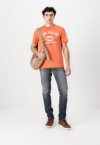 Camiseta de algodón naranja con texto blanco, jeans de mezclilla azul ajustados y zapatillas marrones. Llevando un bolso de hombro beige.