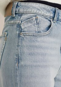 Los jeans de denim azul claro presentan un diseño clásico con costuras visibles, múltiples bolsillos y una textura ligeramente desgastada.