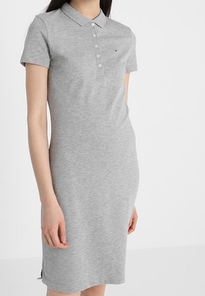 Femme portant une robe polo gris clair à manches courtes avec un col et des boutons, debout devant un fond blanc uni.