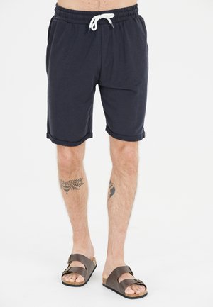 Mørkeblå bomulds shorts med elastisk talje og hvid snor. Har rullede kanter og bæres med brune sandaler.