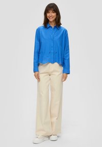 Camicia blu con bottoni, orlo ondulato e dettagli tagliati. Abbinata a pantaloni larghi color crema e sneakers bianche. Sfondo neutro.