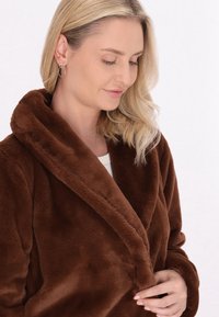 Bruine, plusachtige badjas met een brede kraag en lange mouwen. Soepele textuur, dubbele knoopsluiting en zichtbaar zijzakken.