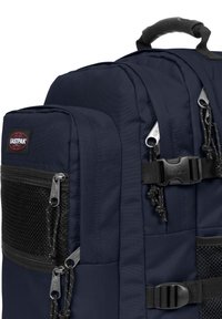 Námorně modrý batoh Eastpak, vyrobený z pevného materiálu, s několika zipovými oddíly, síťovými detaily a nastavitelnými popruhy s kovovými prvky.
