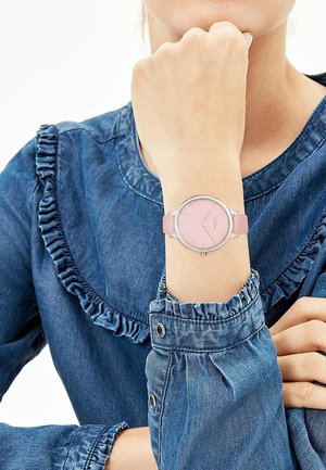 Person wearing a pink-faced wristwatch with a pink strap, dressed in a blue denim top with ruffled details on sleeves and chest.