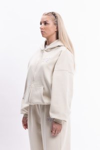 Béžová mikina na zips s veľkou kapucňou a prednými vreckami, vyrobená z mäkkej tkaniny. Má vyšívané detaily a voľné rukávy.