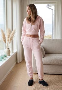Femme en sweat à capuche court rose clair et jogging, les mains dans les poches, debout près de la fenêtre avec vue sur la mer dans un salon lumineux.