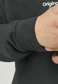 Camisa de manga larga gris oscuro con puños acanalados y un pequeño logo blanco. La tela parece suave y ligeramente elástica.