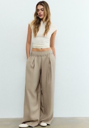 Vrouw in beige broek met wijde pijpen en metalen kettingriem, een kort wit mouwloos topje, staand tegen een effen lichte muur.