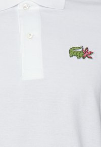 Polo bianco realizzato in tessuto testurizzato, con un coccodrillo ricamato verde e un dettaglio di fiore rosa. Collo con chiusura a tre bottoni.