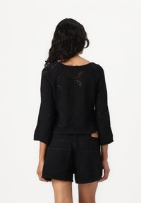 Zwarte gebreide pullover met een gestructureerd bloemenpatroon, voorzien van driekwartmouwen en een brede halslijn, gecombineerd met zwarte shorts.