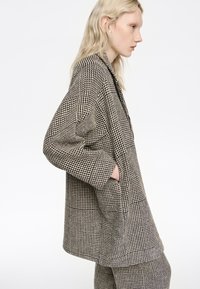 Mujer con cabello largo y rubio que lleva un blazer oversize de pata de gallo en blanco y negro y pantalones a juego, con las manos en los bolsillos, mirando hacia la derecha.