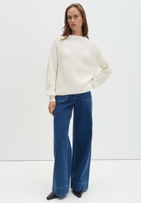 Pull en maille côtelée blanche à col montant, associé à un jean en denim bleu foncé à jambes larges, avec un détail subtil à l'ourlet et des chaussures noires.
