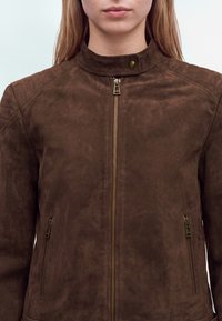 Personne portant une veste en daim marron foncé avec une fermeture éclair à l'avant et des poches latérales zippées, montrée des épaules jusqu'au milieu du torse.