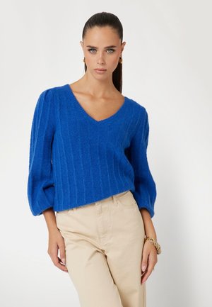 Maglione blu a V in maglia con texture a coste e maniche a palloncino, abbinato a pantaloni beige. Il materiale sembra morbido e accogliente.