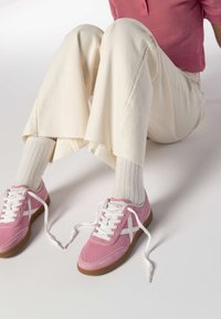 Baskets en daim rose avec des lacets blancs et un design blanc sur le côté, associées à un jean crème effiloché et des chaussettes blanches côtelées.